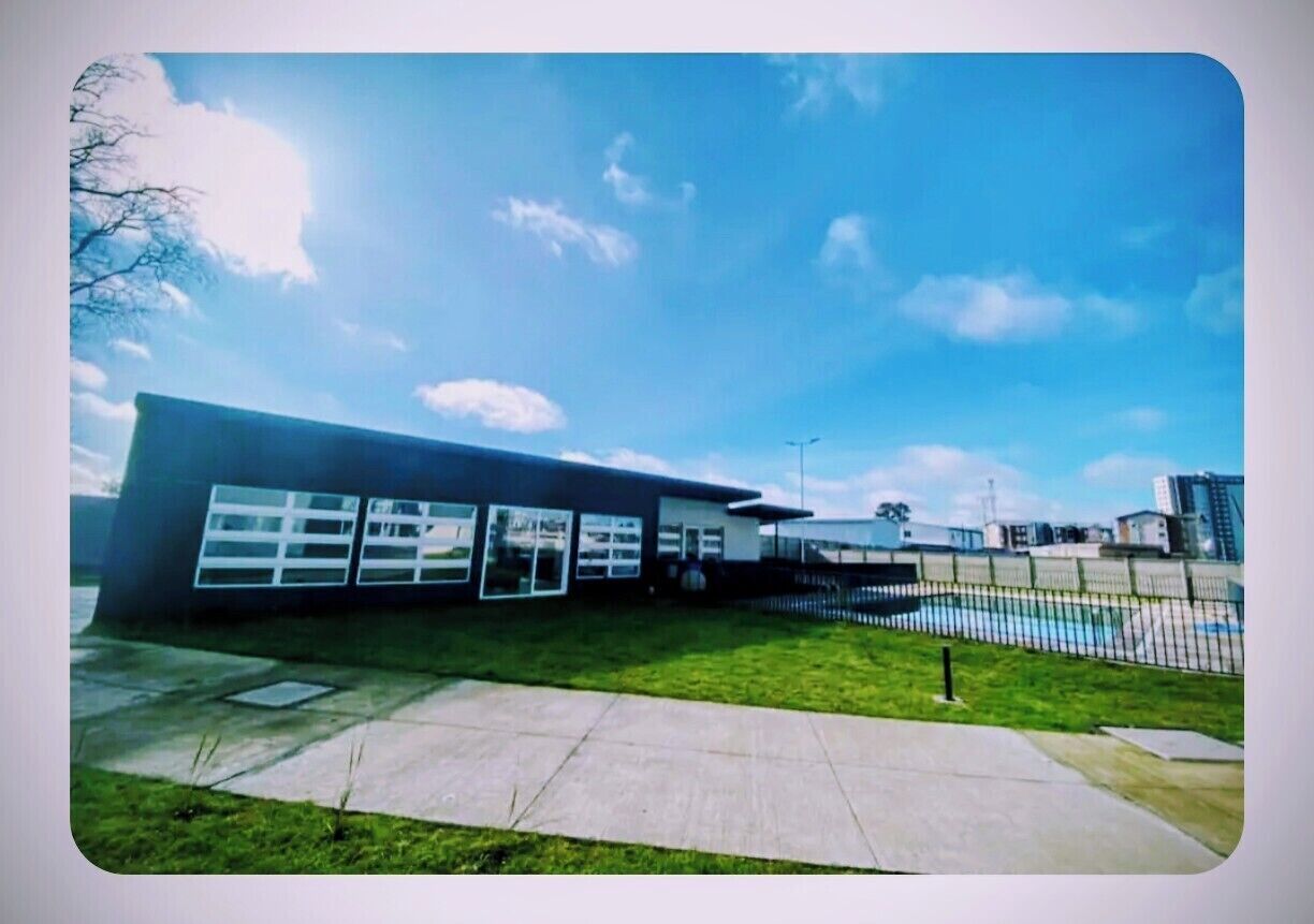 A modern building with large windows and a flat roof, surrounded by a fenced area and a clear sky.