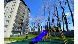 Une aire de jeux avec un toboggan bleu et des balançoires, entourée d’arbres et d’un bâtiment.