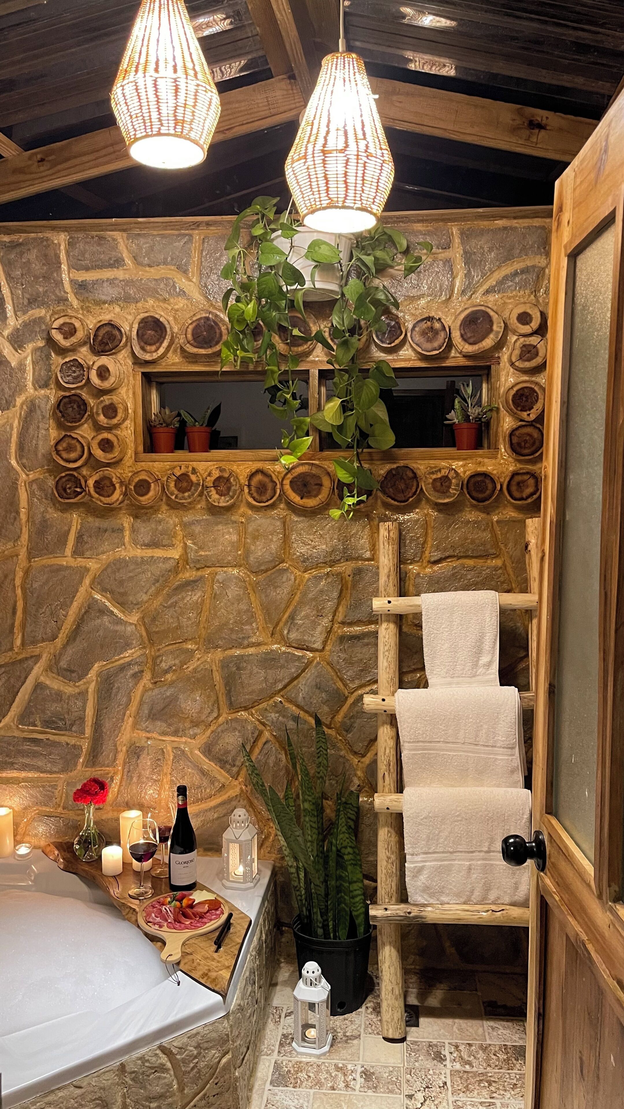 Een badkamer met stenen muren, een houten ladder, een hangplant, kaarsen en een fles wijn.
