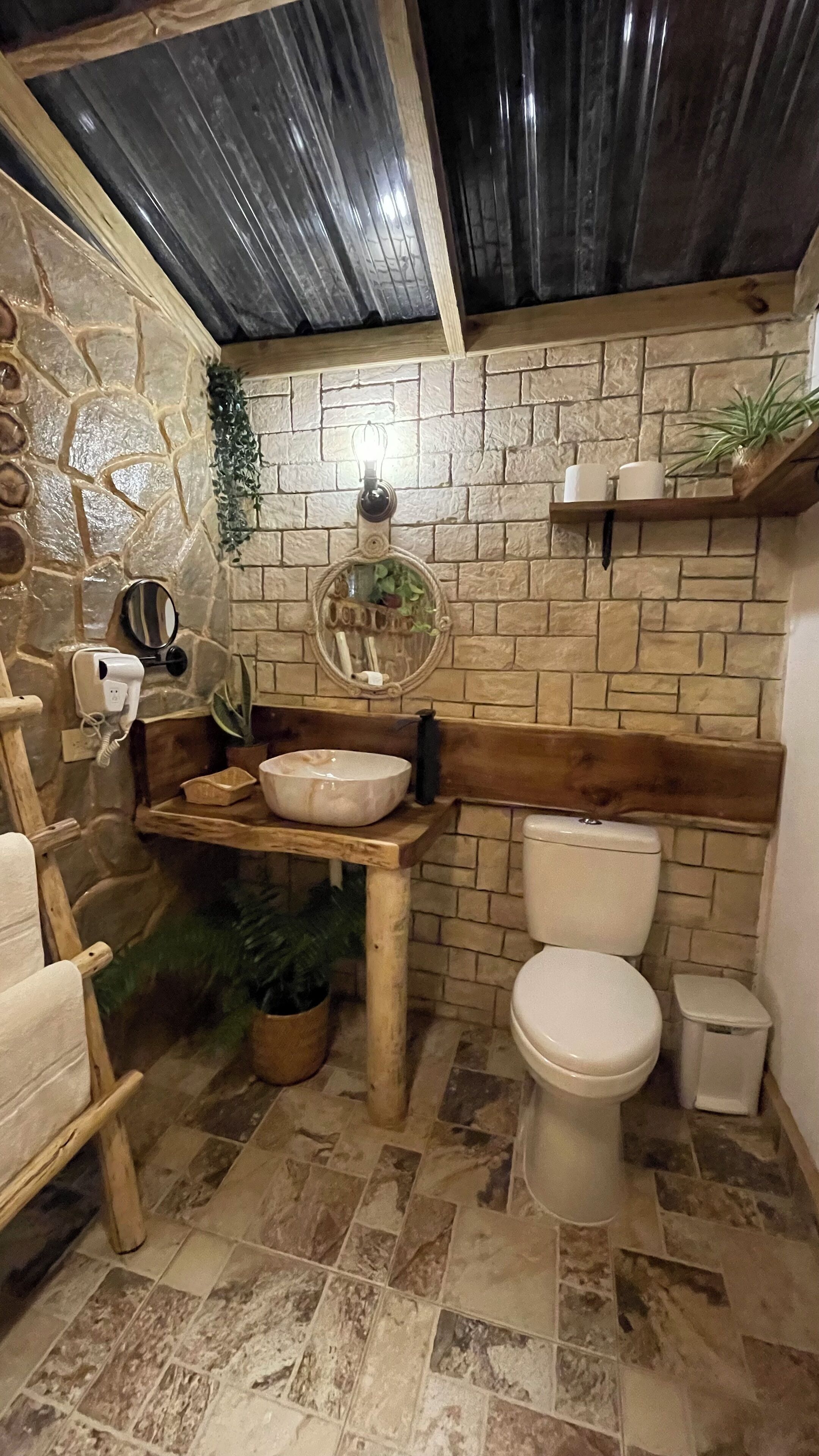 A bathroom with a stone wall, wooden bench, sink, toilet, and a mirror.