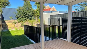 Una terraza de madera con cerca de metal negro, zona cubierta de césped y un edificio con tejado rojo al fondo.