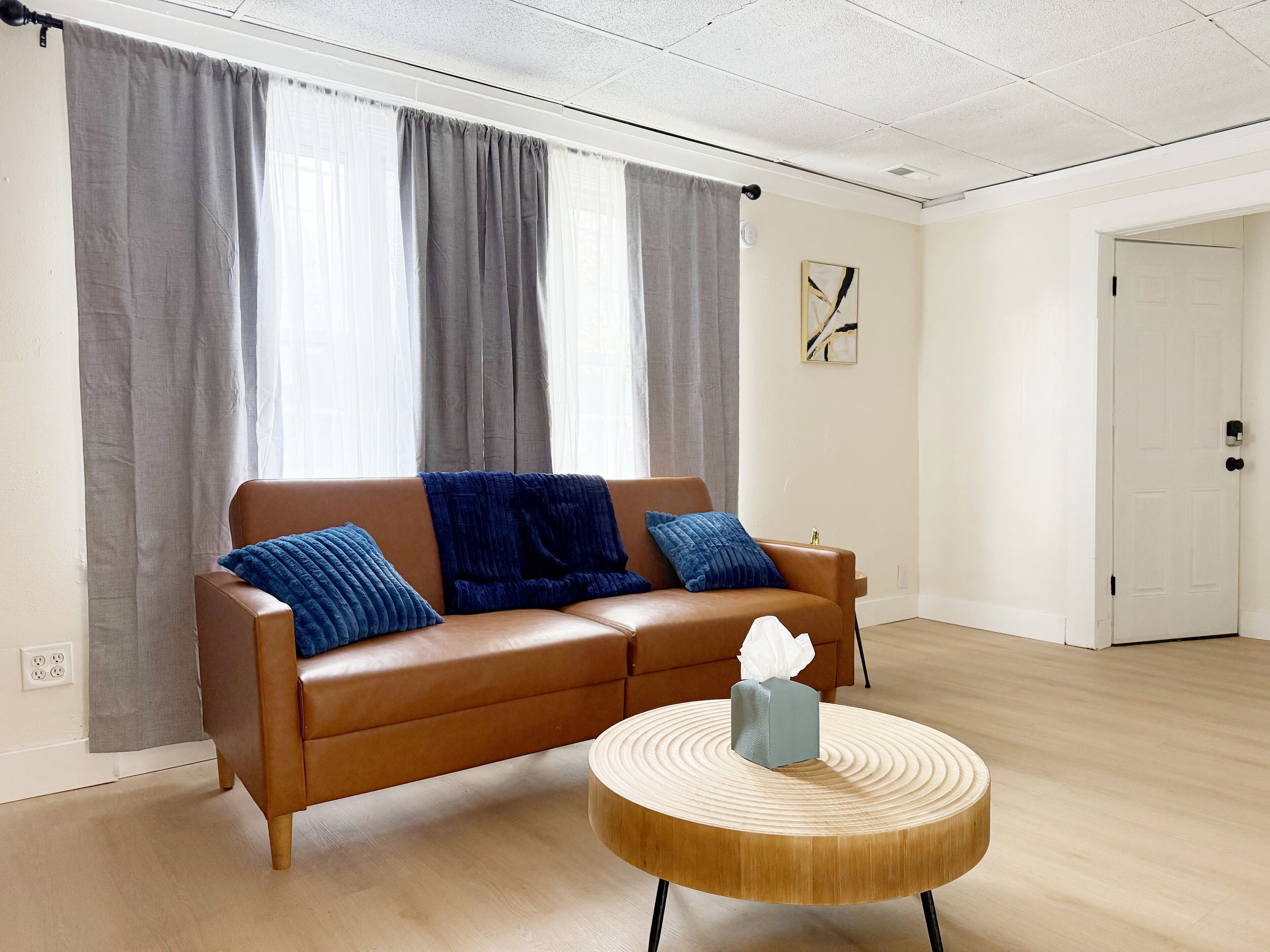 A spacious living room with a brown leather sofa, three blue cushions, a round wooden coffee table, and a white door in the background.