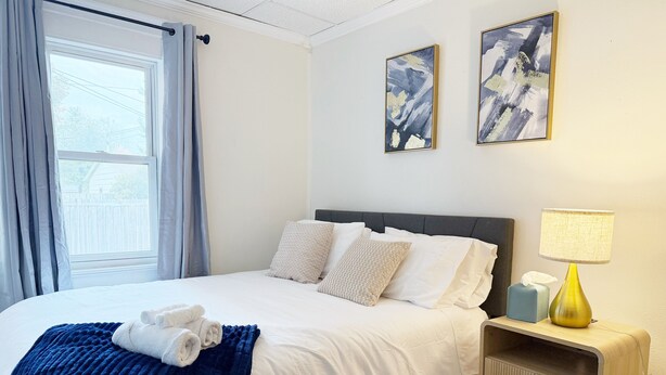 Una cama bien tendida con sábanas blancas y una manta azul, una mesita de noche con una lámpara, y dos cuadros abstractos en la pared.
