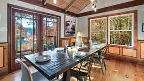 Zona pranzo con tavolo in legno apparecchiato per quattro, ampie finestre con vista sugli alberi e soffitto in legno con travi a vista.