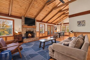 A spacious living area with a stone fireplace, wooden ceiling beams, and large windows offering views of trees. - FreeDollywoodTix+Pool+Secluded+Modern+HotTub (Sevierville)