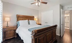 A bedroom with a wooden bed, two bedside tables, a ceiling fan, and a door leading to another room. - Dallas Home for Extended Stays: 3BR, Pool, Workspace and Garage (Dallas)