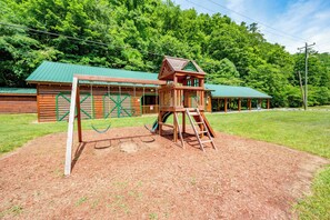 Eine Spielkonstruktion aus Holz mit einer Leiter und Schaukeln in einer grünen Wiese, im Hintergrund eine Blockhütte.