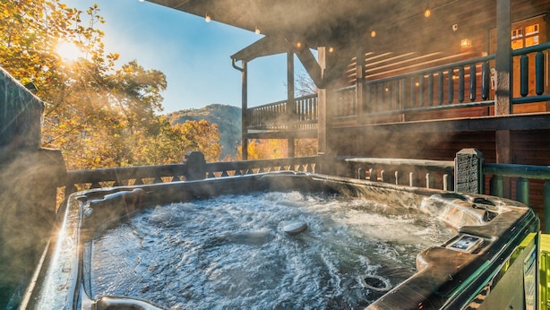A hot tub with steam rising, surrounded by a wooden deck and a covered area with string lights. - Free Dollywood Tickets, Sauna, Hot Tub, Secluded Modern Cabin, Dogs OK (Sevierville)