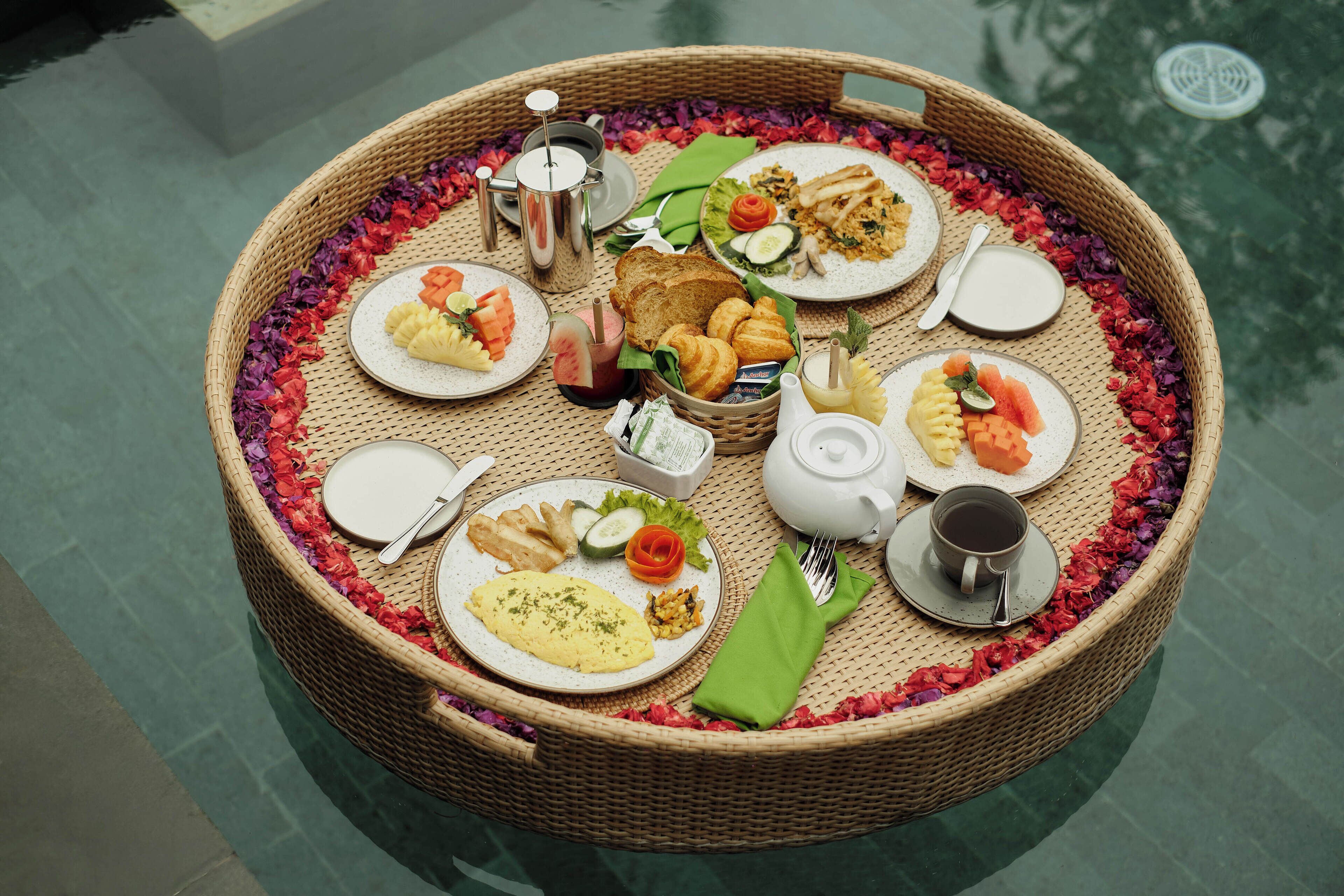 Una bandeja de mimbre con alimentos para el desayuno, como tostadas, huevos y fruta, sobre una mesa de vidrio junto a una piscina.
