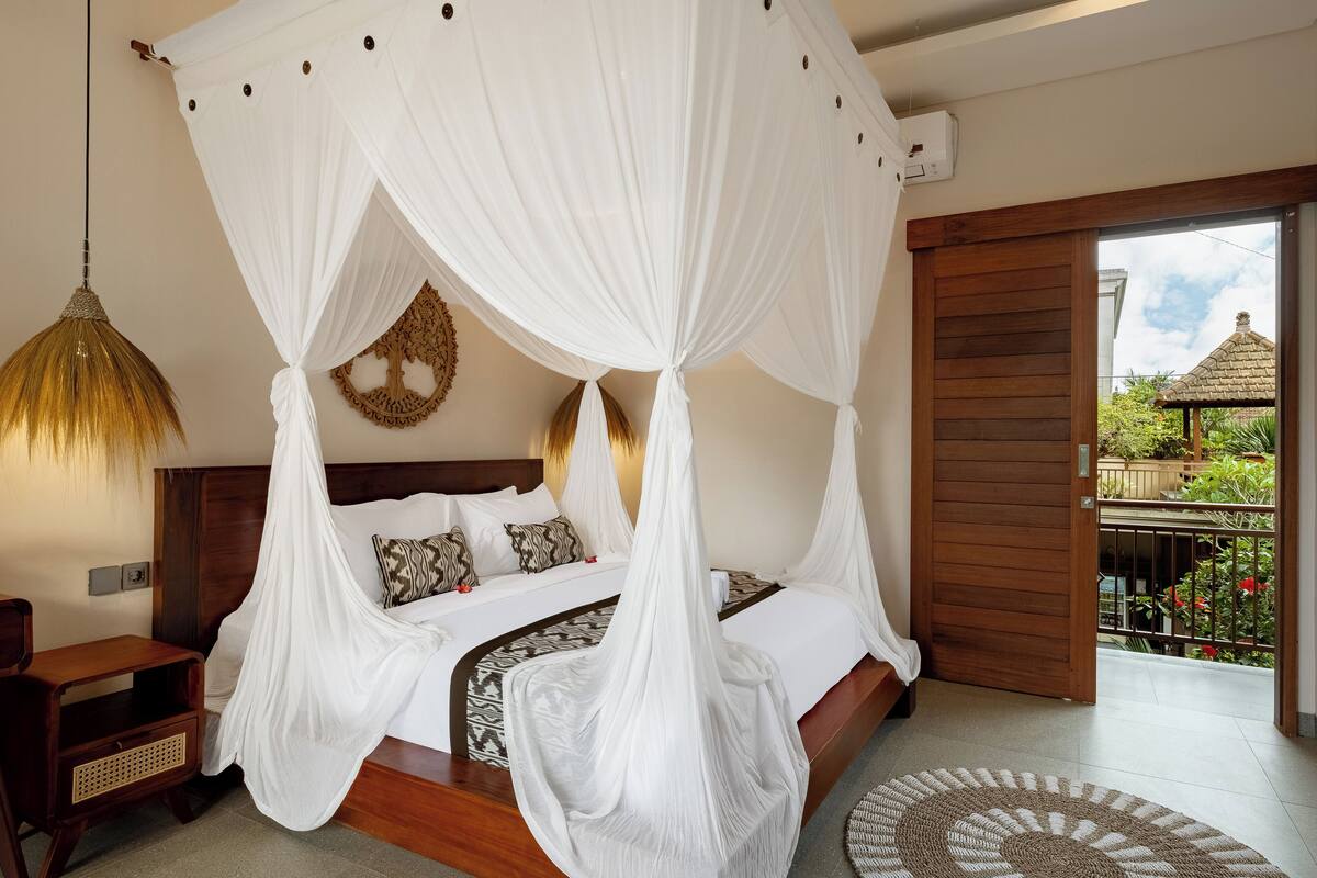 A bedroom with a four-poster bed, a bedside table, a hanging lamp, and a view of a garden through an open door.
