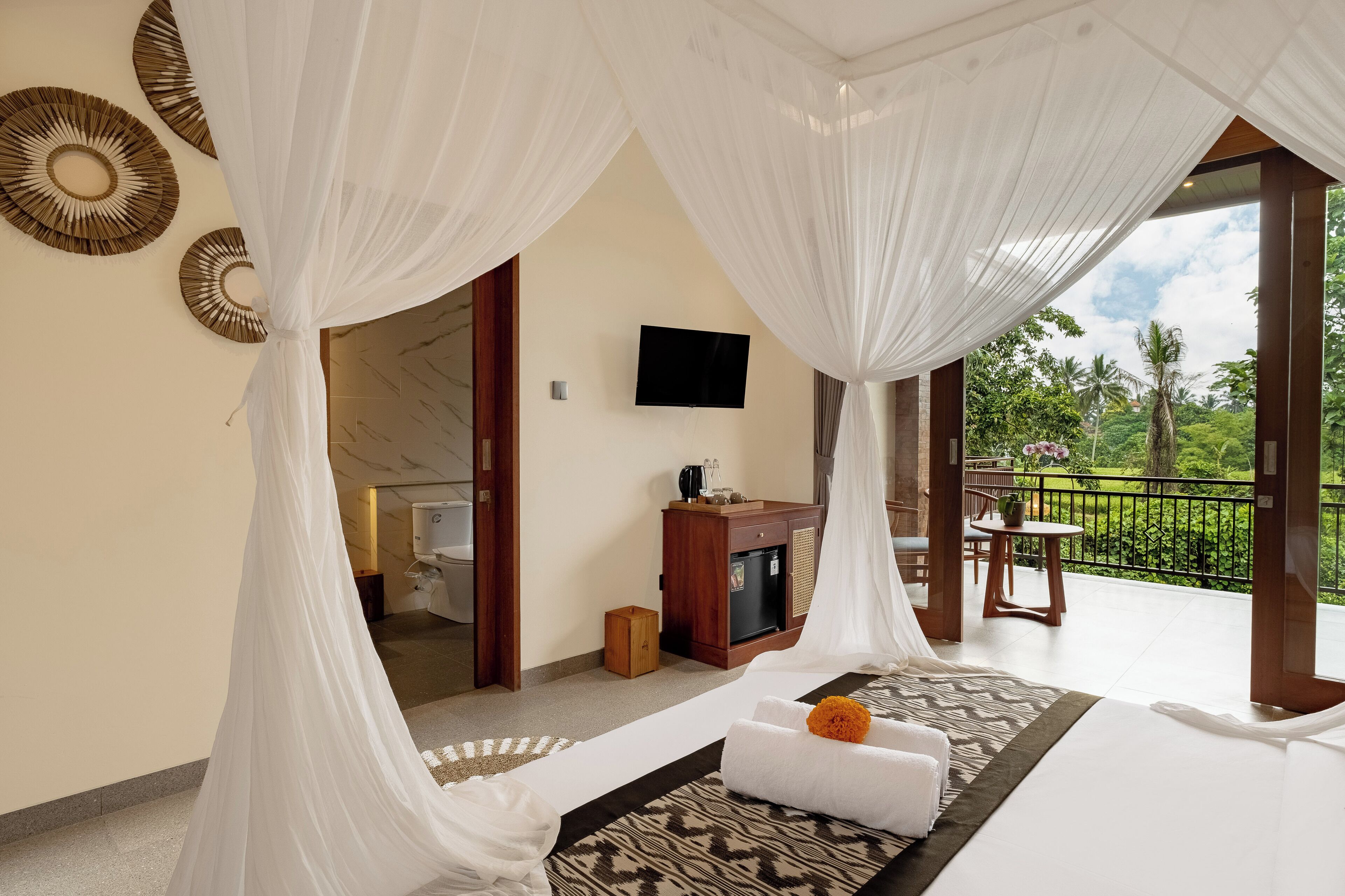 A bedroom with a bed, a TV, a small table, and a balcony with a view of greenery.
