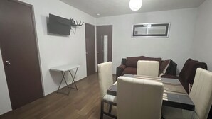 A room with a brown door, a white wall-mounted TV, a window, a brown sofa, a white sofa, and a dining table with chairs.