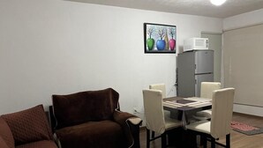 A living room with a brown sofa, a dining table with chairs, a gray refrigerator, and a framed picture on the wall.