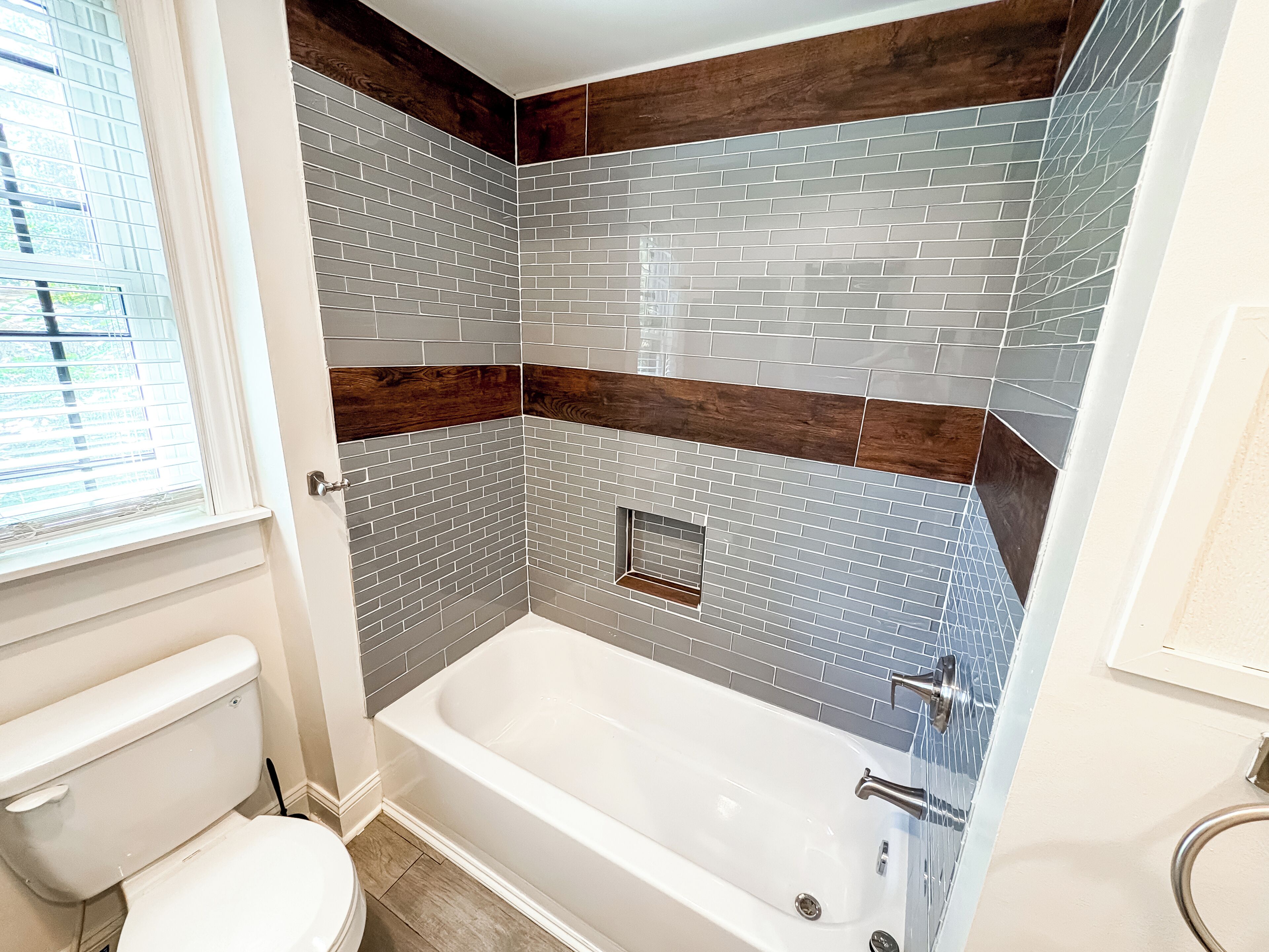 A bathroom with a white bathtub, grey and brown tiled walls, and a window with blinds.