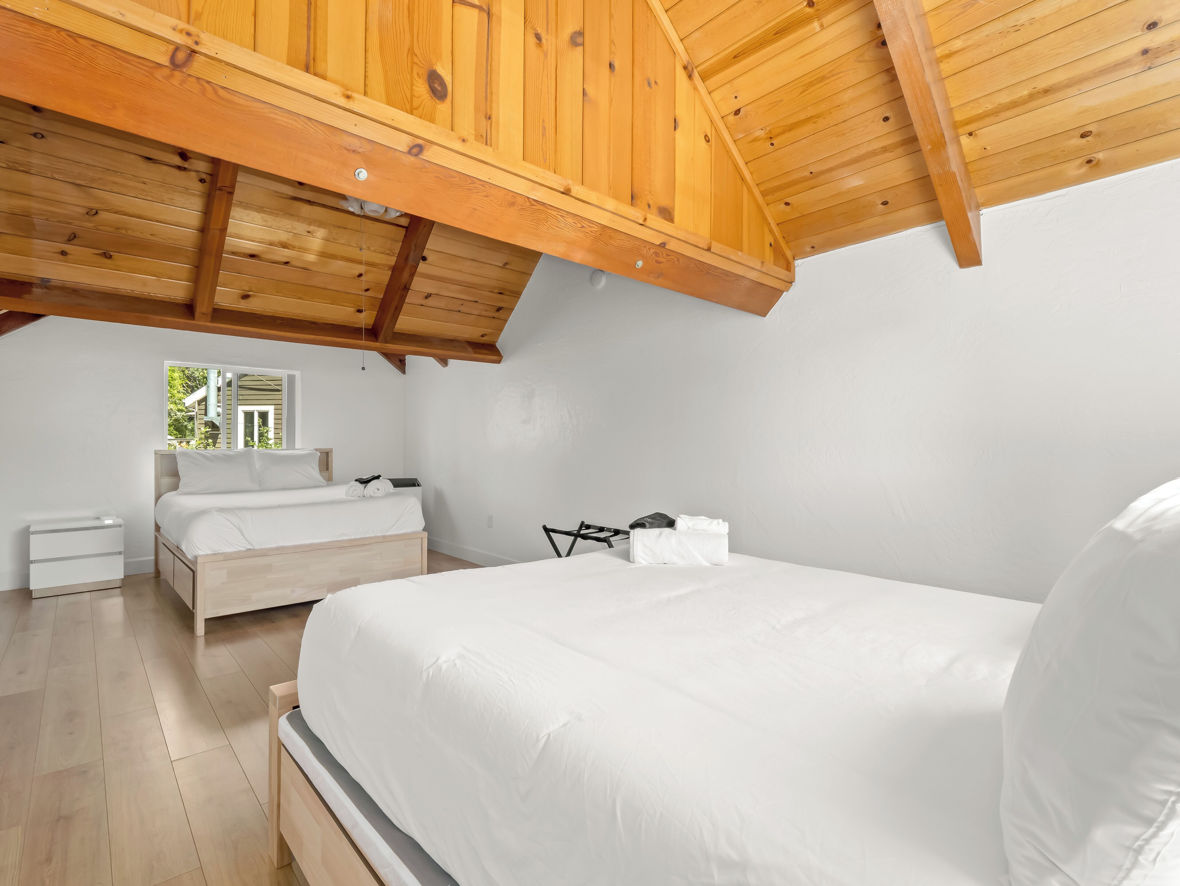 Een slaapkamer met een houten plafond, een bed, een nachtkastje en een raam met uitzicht op groen.