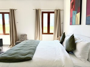 Une chambre avec un grand lit, des rideaux beiges, une vue sur l’extérieur et un tableau accroché au mur.