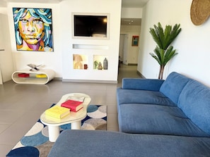 Un salon moderne avec un canapé bleu, une table basse en verre sur laquelle sont posés des livres, un téléviseur fixé au mur et une plante décorative.