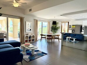 A spacious living area with a sofa, a coffee table, and a dining table with chairs. There is a large window with curtains, a ceiling fan, and a kitchen area with modern appliances. - NEW! Modern and spacious 2-bedroom by the marina in Porto Cupecoy (Sint Maarten)