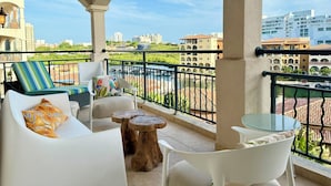 A balcony with white furniture, a glass table, and a view of a pool and buildings. - NEW ! Elegant and modern 2-Bedroom, with marina and garden view in Porto Cupecoy (Sint Maarten)