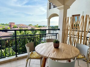 A balcony with a round wooden table and two chairs, a potted plant, and a view of buildings and greenery. - NEW ! Elegant and modern 2-Bedroom, with marina and garden view in Porto Cupecoy (Sint Maarten)