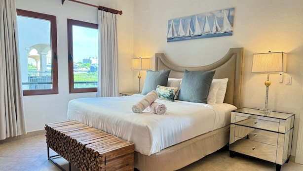 A hotel room with a large bed, two bedside lamps, a wooden bench, and a view of the outdoors. - NEW ! Elegant and modern 2-Bedroom, with marina and garden view in Porto Cupecoy (Sint Maarten)
