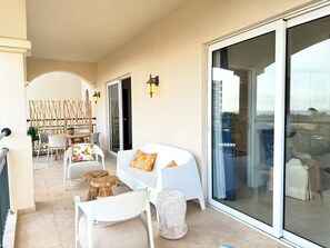A balcony with a white sofa, wooden stools, and a dining area with chairs and a table. - NEW ! Elegant and modern 2-Bedroom, with marina and garden view in Porto Cupecoy (Sint Maarten)
