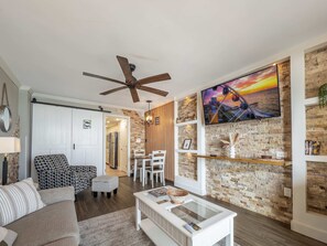 A modern living room with a ceiling fan, a dining area with white chairs, a kitchen in the background, and a brick wall with a mounted TV. - Caravelle 937 (Myrtle Beach)