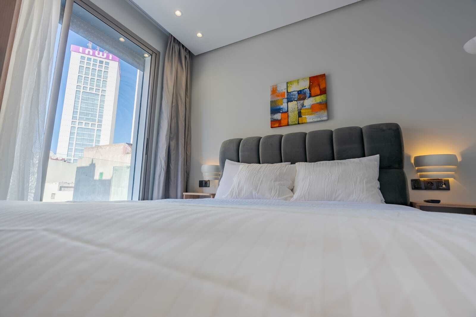 A hotel room with a large bed, a view of a cityscape through a window, and a modern abstract painting on the wall.