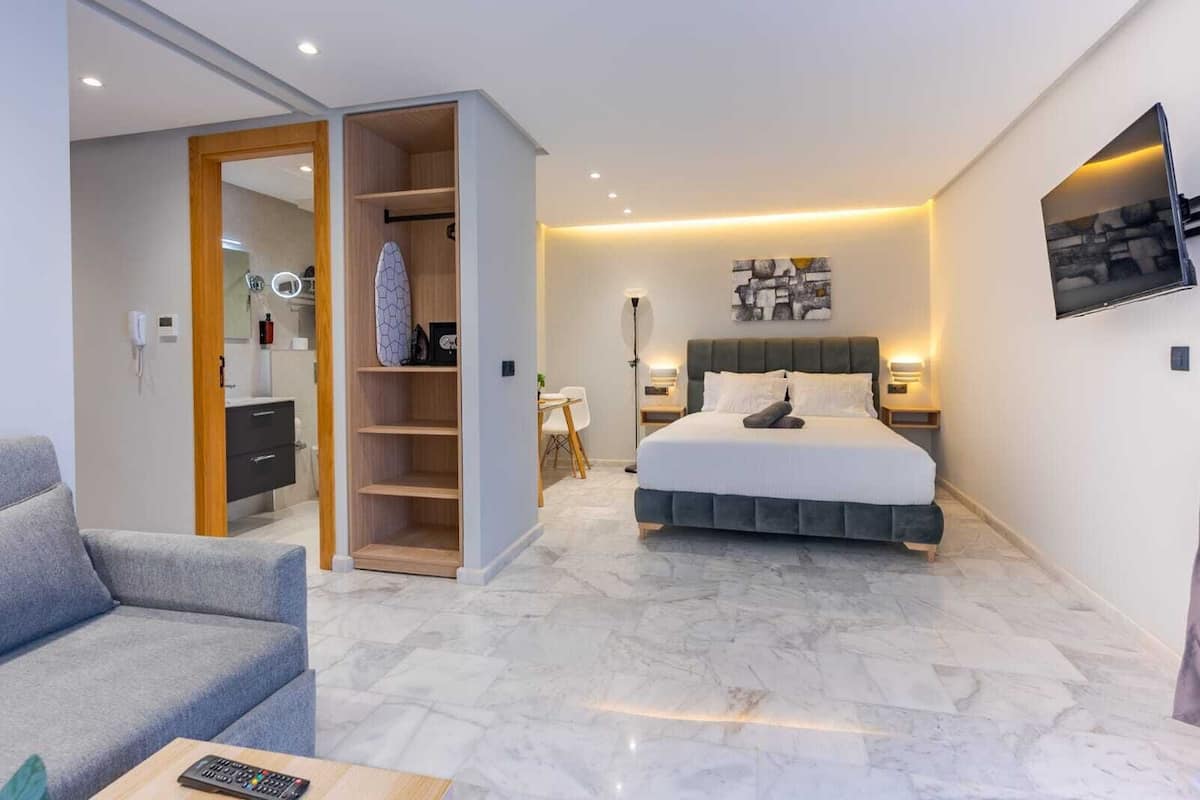 Een moderne hotelkamer met een bed, een zithoek met een bank, een inloopkast en een badkamer met spiegel en handdoekhouder.