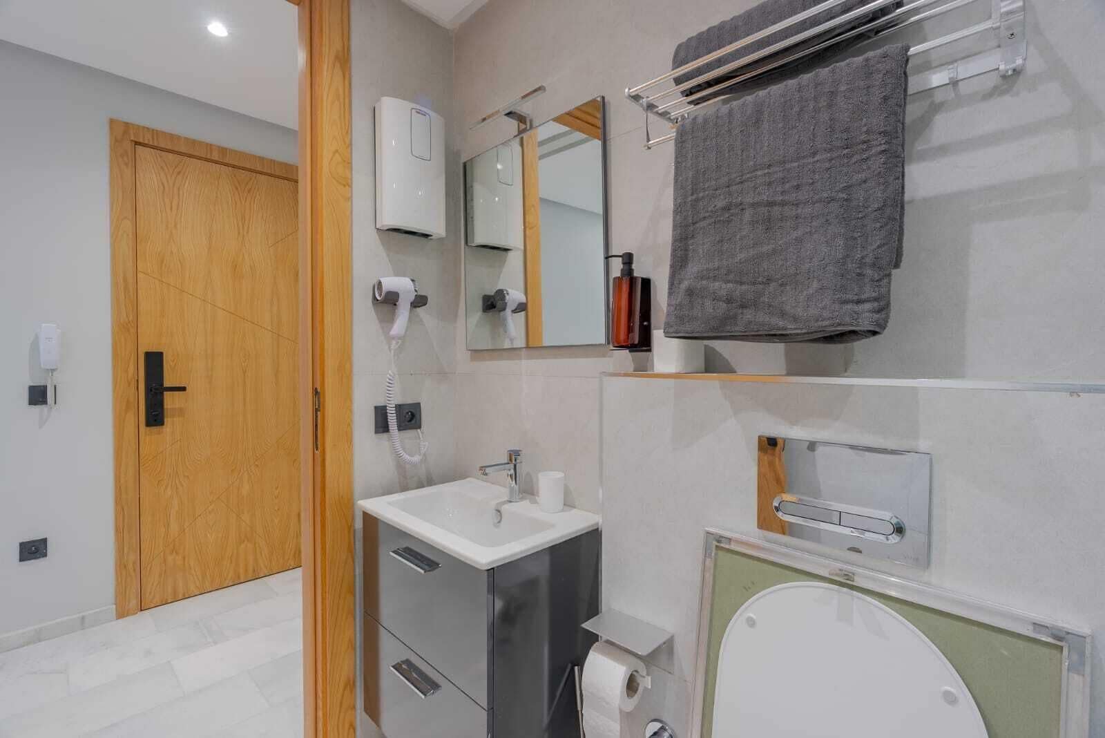 A modern bathroom with a wooden door, a white sink, a mirror, a towel rack, and a toilet.