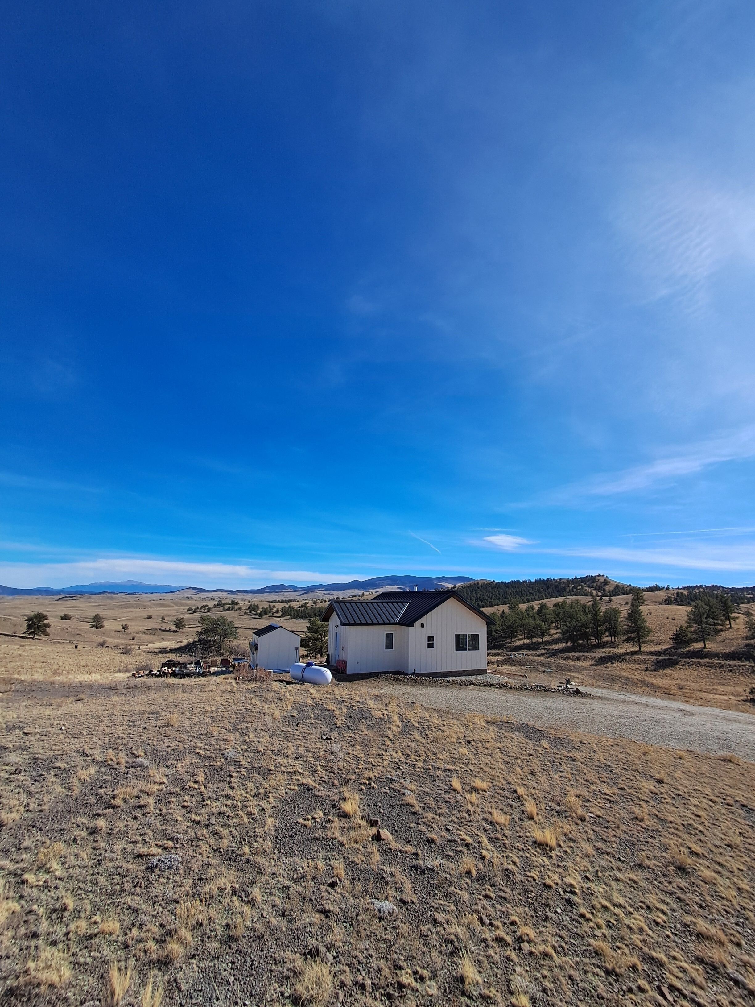 A single-story house with a black roof and white walls, surrounded by a dry, grassy landscape with a clear blue sky.