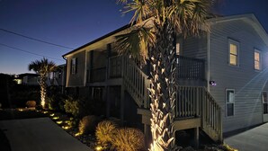 A two-story house with a balcony, a palm tree, and a well-lit garden area. - Soundfront Escape • Private Heated Pool • Dock • Pet Friendly (Emerald Isle)