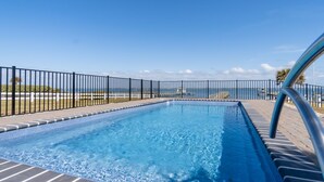A rectangular swimming pool with a clear blue water surface, surrounded by a black metal fence, and a view of the ocean in the background. - Soundfront Escape • Private Heated Pool • Dock • Pet Friendly (Emerald Isle)