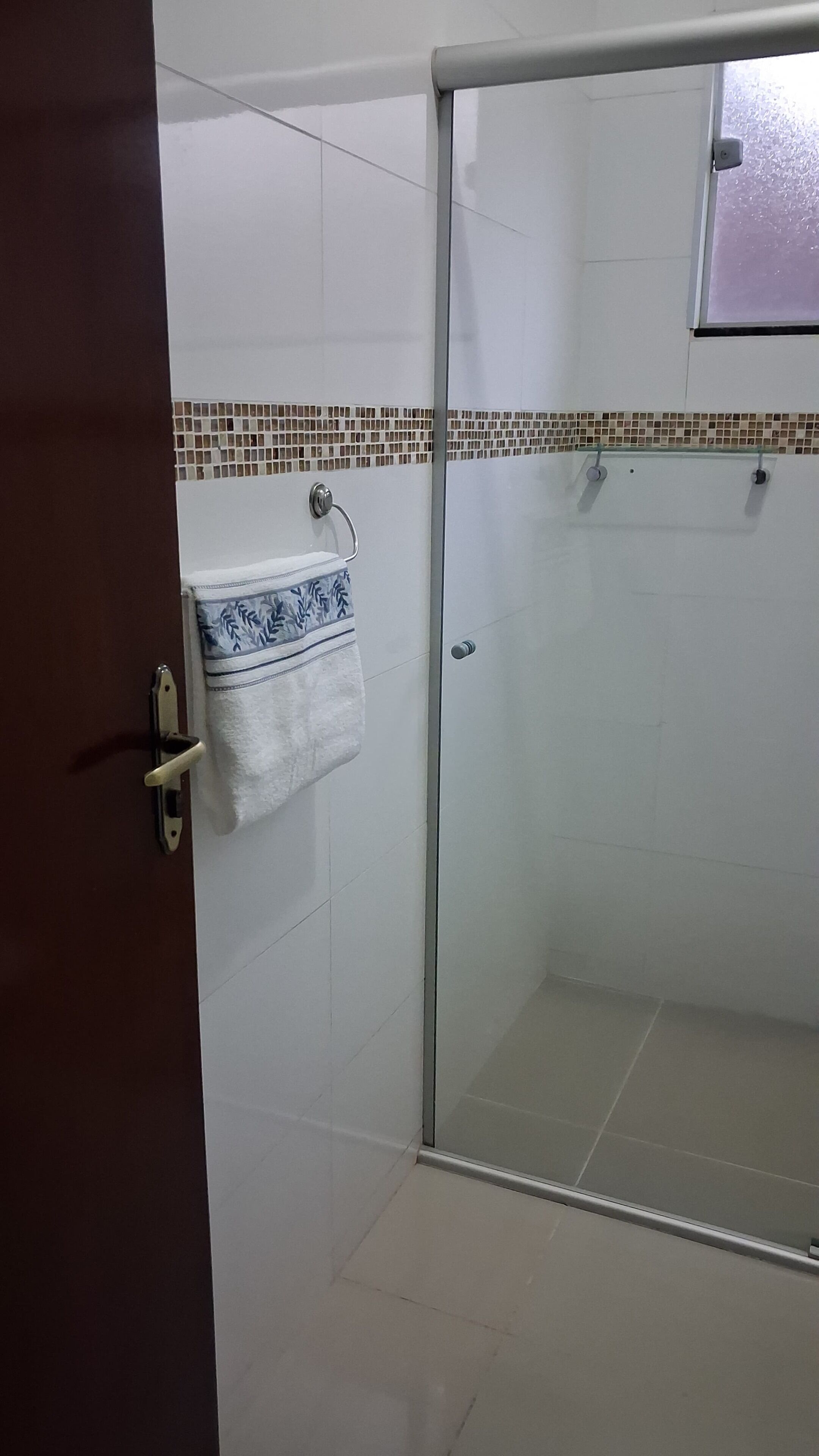Un baño con mampara de vidrio, paredes de azulejos blancos, una toalla estampada colgada en la puerta y un pequeño estante sobre el lavamanos.