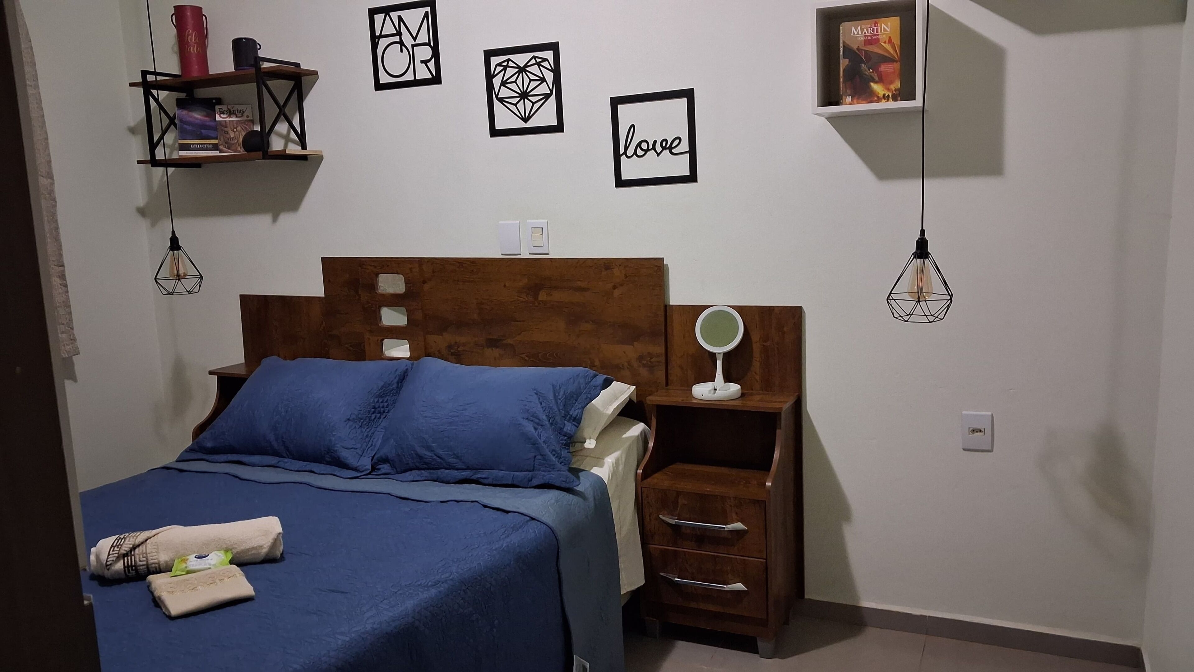 Une chambre à coucher avec une tête de lit en bois, une couvre-lit bleue, une table de chevet avec un miroir et une étagère avec des livres et une tasse rouge.