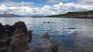 A rocky shoreline with clear water and a person swimming in the distance.