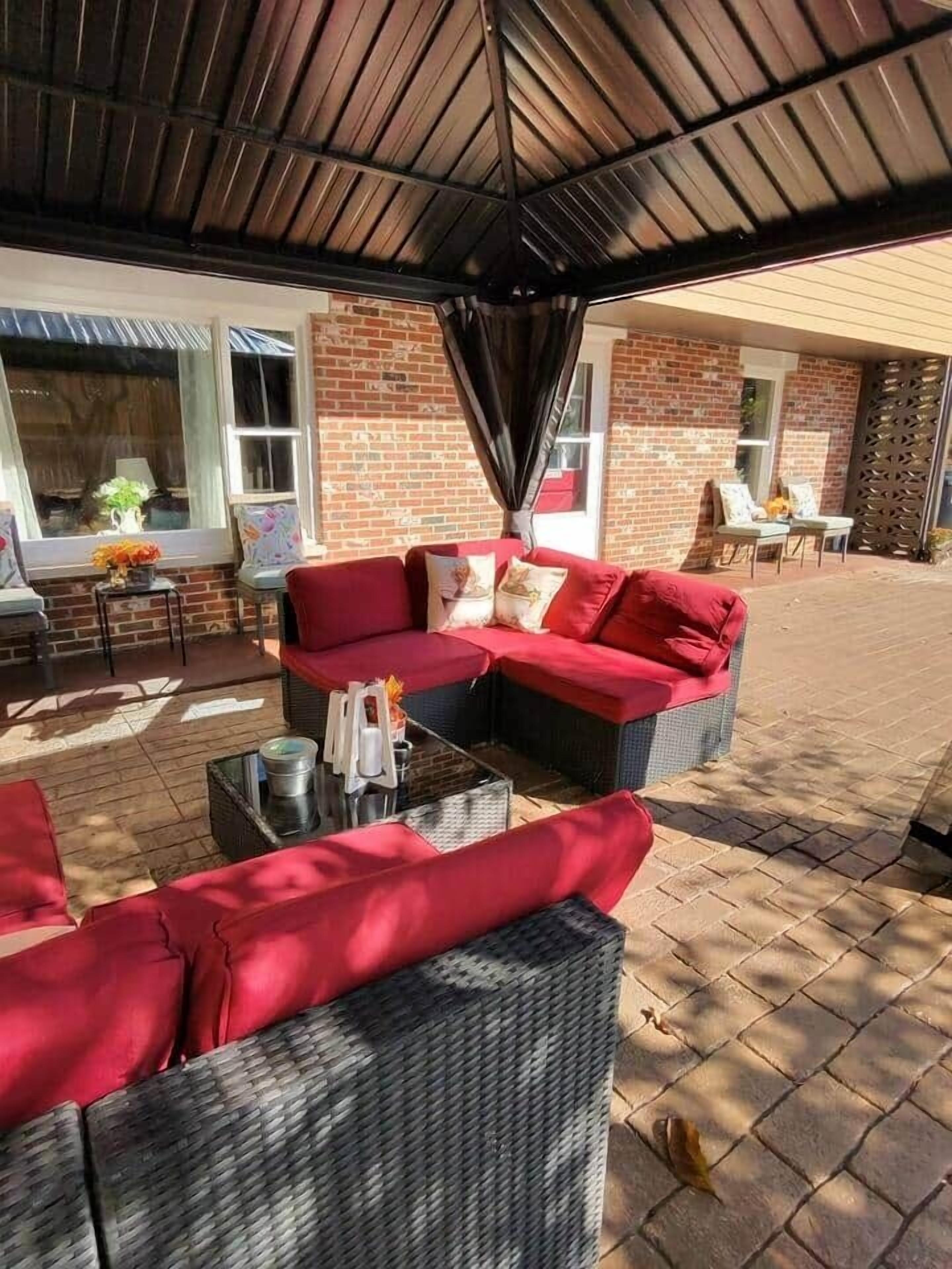 A covered outdoor seating area with red sofas and wicker tables, brick walls, and a wooden ceiling.