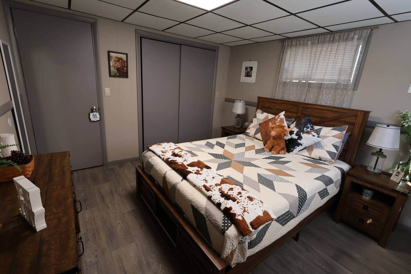 Een slaapkamer met een houten bed, nachtkastje met een lamp, een dressoir en een raam met jaloezieën.
