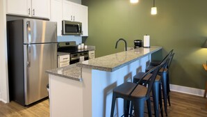 A modern kitchen with a stainless steel refrigerator, a microwave, a stove, and a sink. There are bar stools and a light fixture above the counter. - Retro 1 Bedroom, King Bed, Large Living Area, Parking, Gym, Close to UAB (Birmingham)