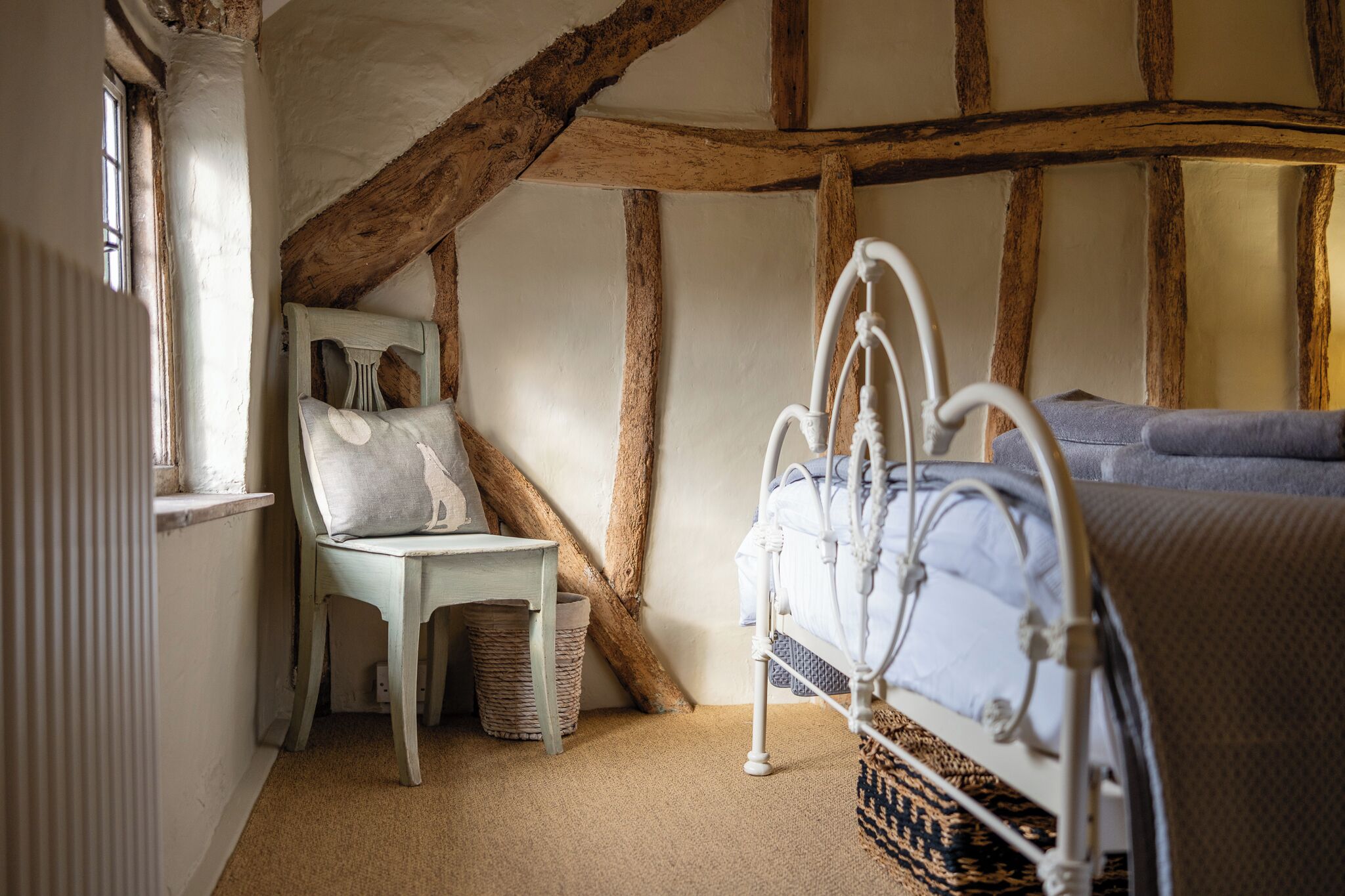 A bedroom with a sloped ceiling, a bed with a white metal frame, a chair with a cushion, and a window with a radiator.