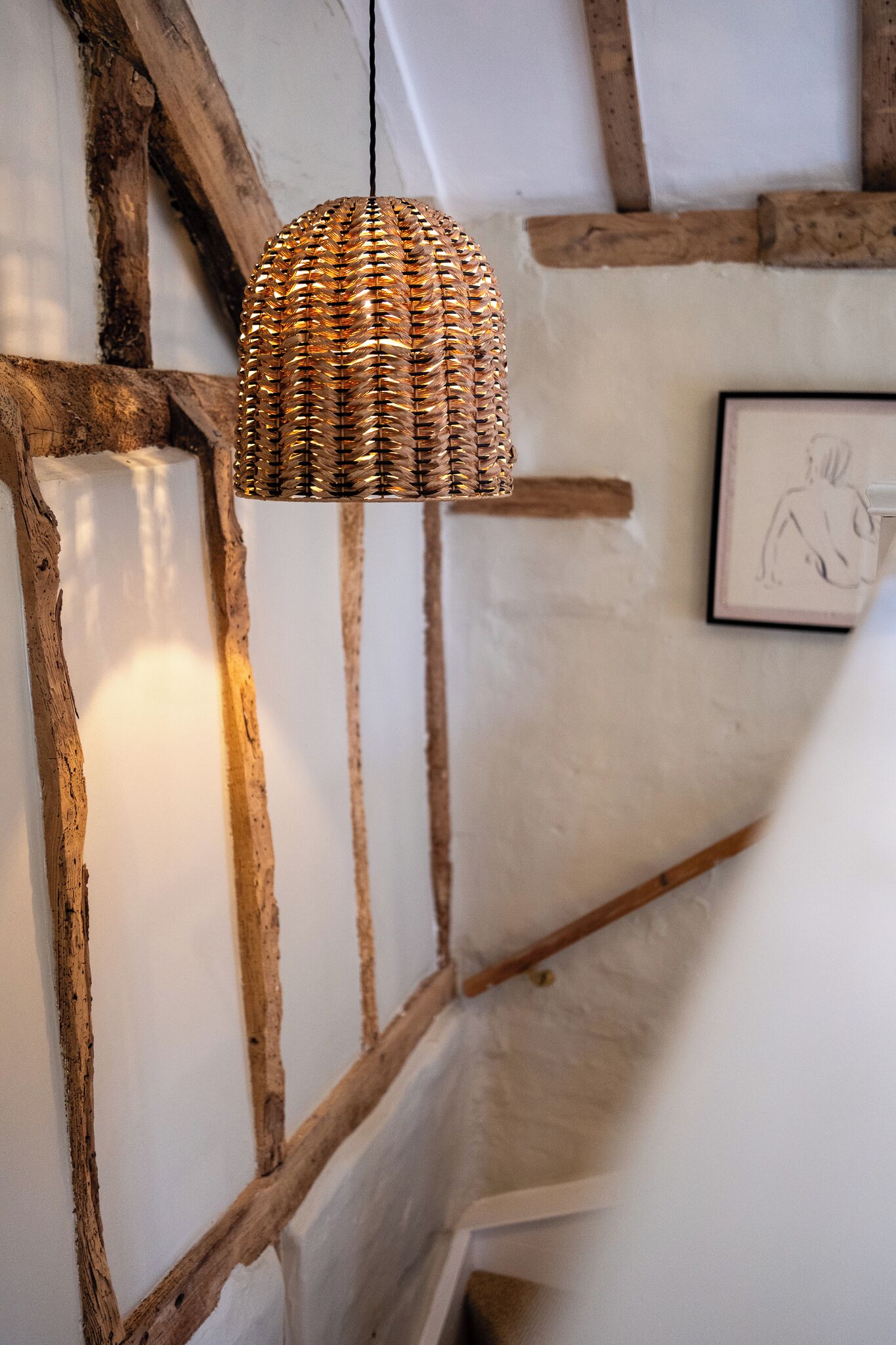 A wicker pendant light hanging from a ceiling with exposed wooden beams and a framed drawing on the wall.