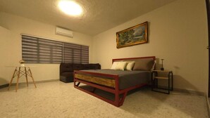 Une chambre à coucher avec un lit, une fenêtre avec des stores, un tableau au mur et une petite table avec une lampe.