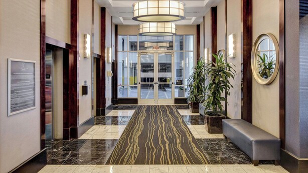 Un vestíbulo de hotel moderno con piso de mármol, un banco, plantas en macetas y un amplio ventanal que da al área de recepción.