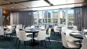Un comedor moderno con mesas dispuestas para comensales, amplias ventanas con vistas a la ciudad y una alfombra estampada.