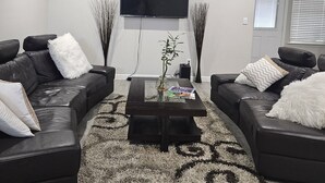 Ein modernes Wohnzimmer mit zwei Ledersofas, einem Glastisch, einem flachbildfernseher an der Wand, einem Deckenventilator und einem gemusterten Teppich.