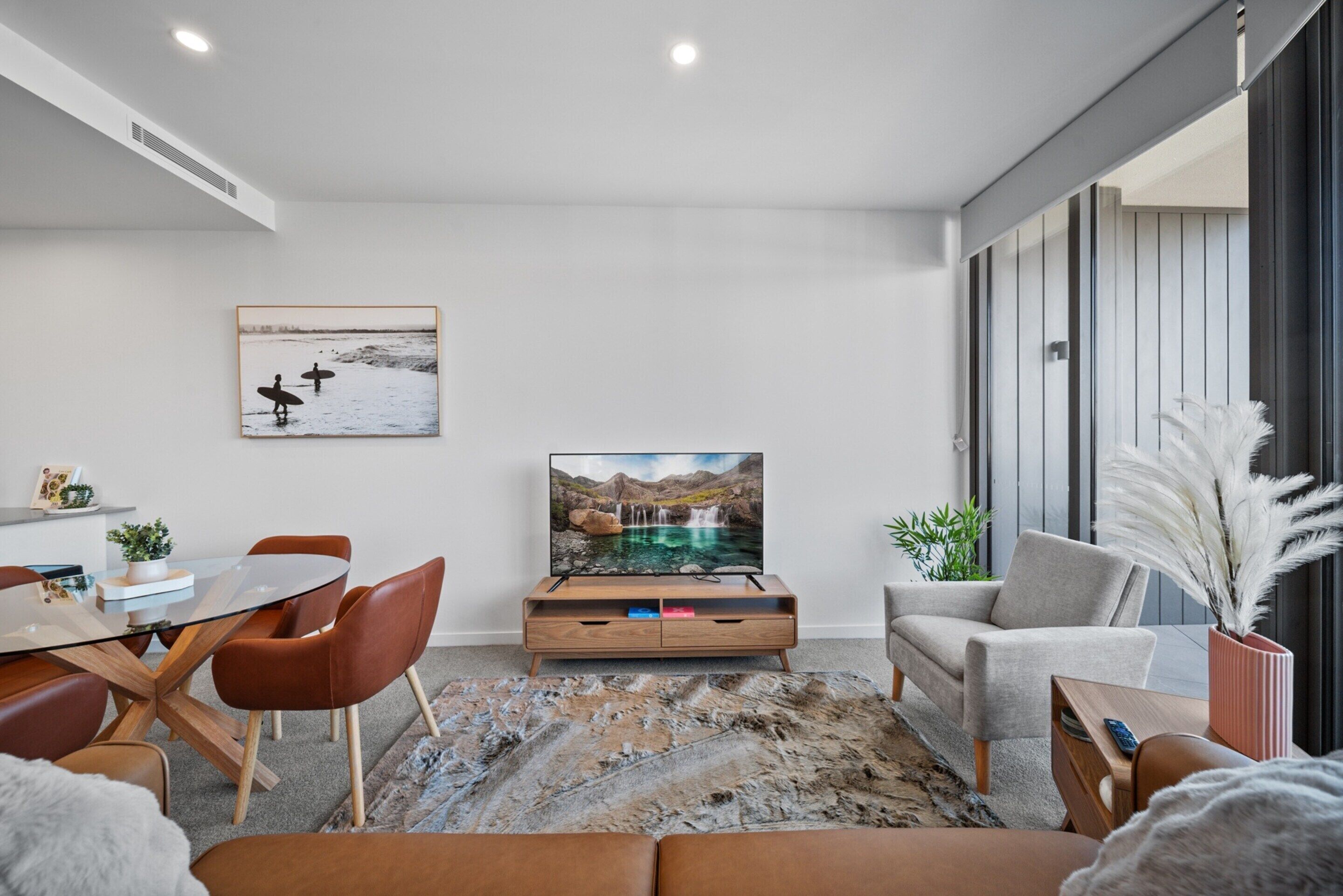 A modern living room with a glass coffee table, brown leather sofa, and a flat-screen TV on a wooden stand.