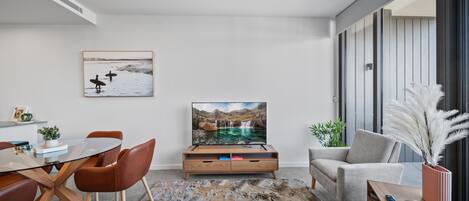 A modern living room with a glass coffee table, brown leather sofa, and a flat-screen TV on a wooden stand.