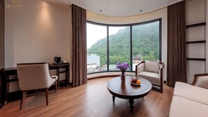 Ein Hotelzimmer mit einem großen, gebogenen Fenster, einem runden Holztisch mit Blumenvase, zwei Stühlen, einem Schreibtisch mit Computer und einer Couch.