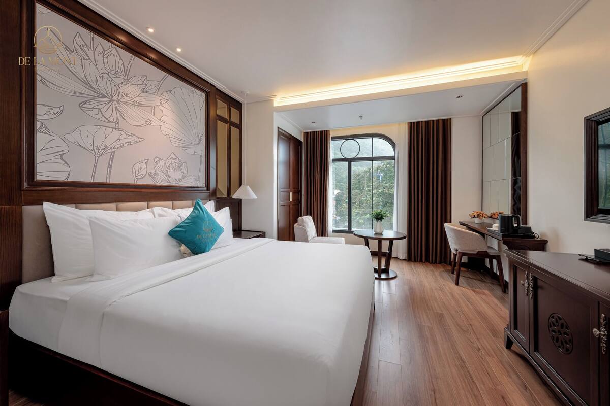 Une chambre d’hôtel avec un grand lit, un bureau, une chaise, une petite table, un miroir et une fenêtre avec des rideaux.