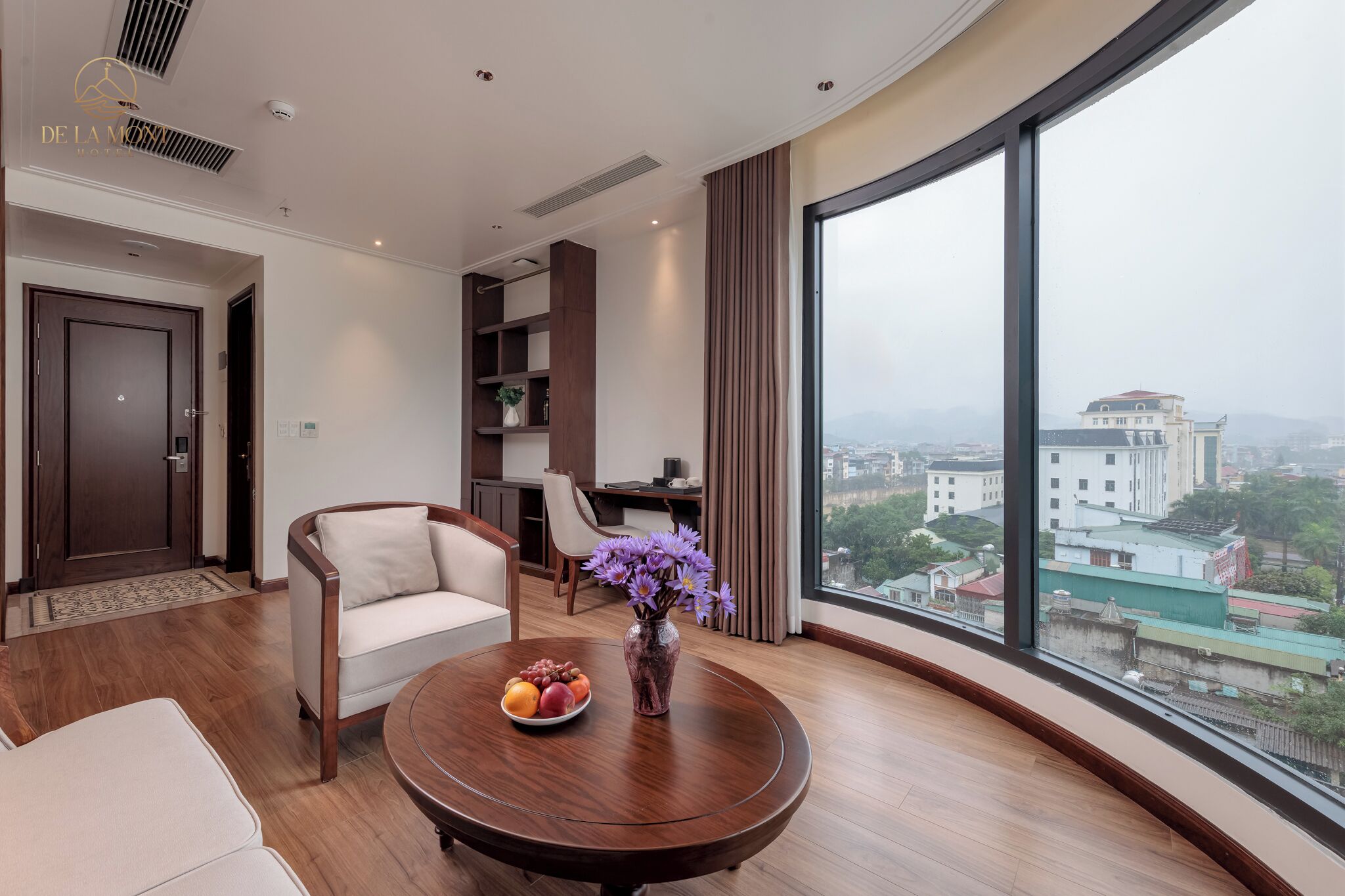 A modern hotel room with a large window, a round wooden table with fruit and flowers, a sofa, and a desk.
